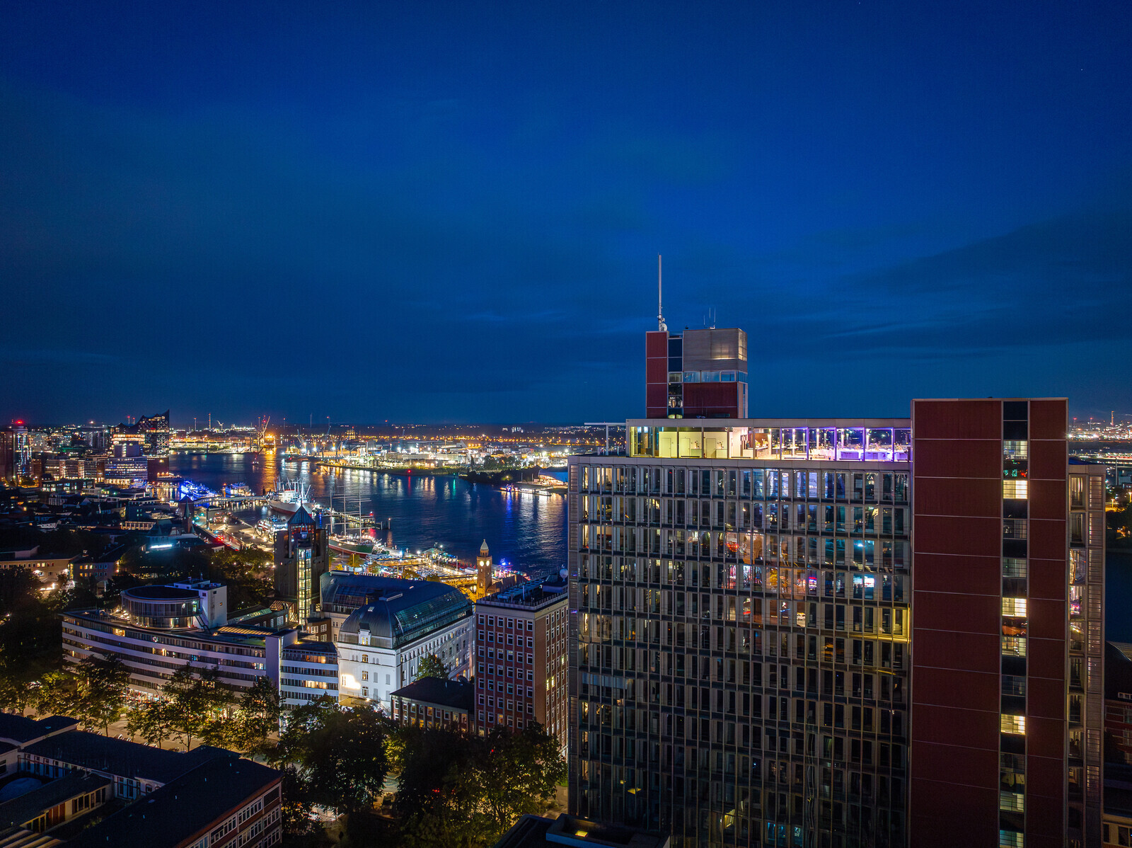 Panoramablick über Hamburg bei Nacht mit Elbe, Hafenlichtern und einem Hochhaus mit beleuchteter Eventlocation auf dem Dach.
