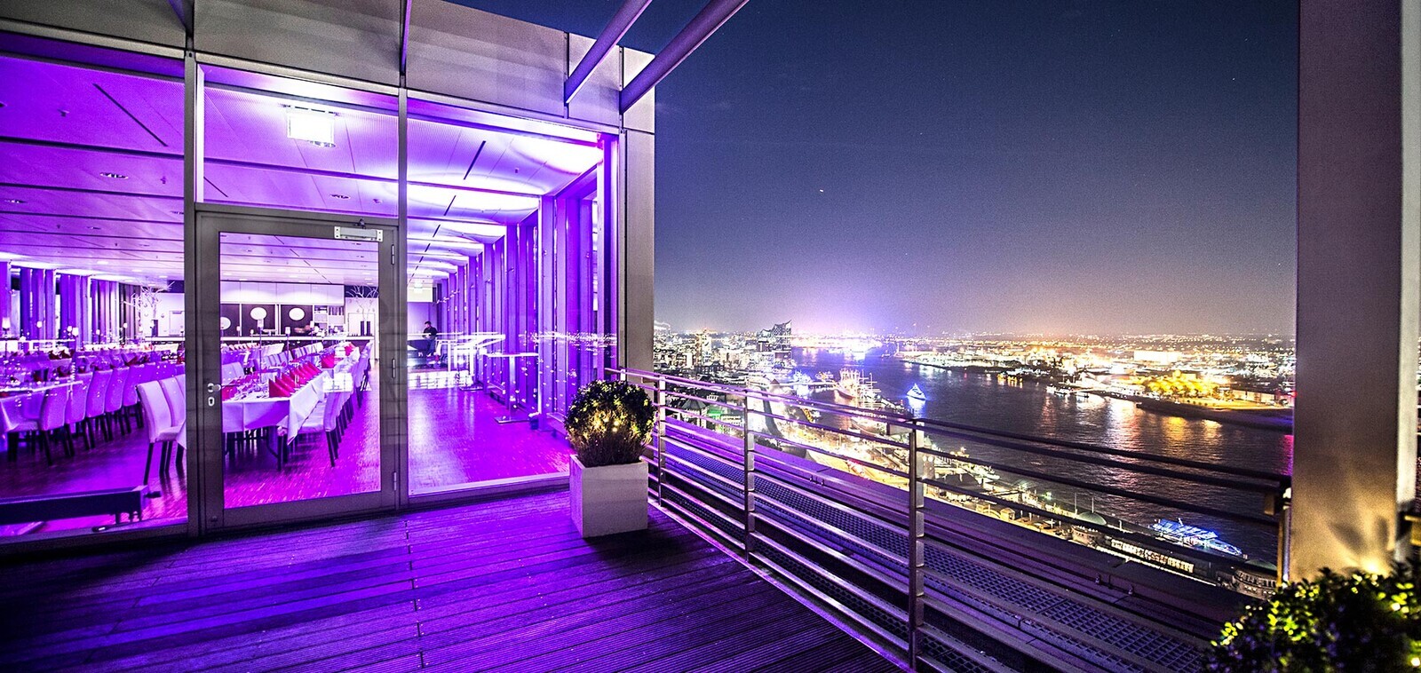 Dachterrasse des Penthouse Elb-Panorama bei Nacht mit violett beleuchtetem Innenraum, bodentiefen Fenstern und Blick über Elbe, Hafen und Hamburger Skyline.